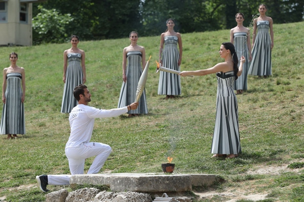 扮演最高女祭司的演員瑪麗·米娜（前右）點燃第一棒火炬手——希臘運動員、東京奧運會男子賽艇項目金牌得主斯特凡諾斯·恩圖斯科斯手中的火炬。新華社記者 趙丁喆 攝